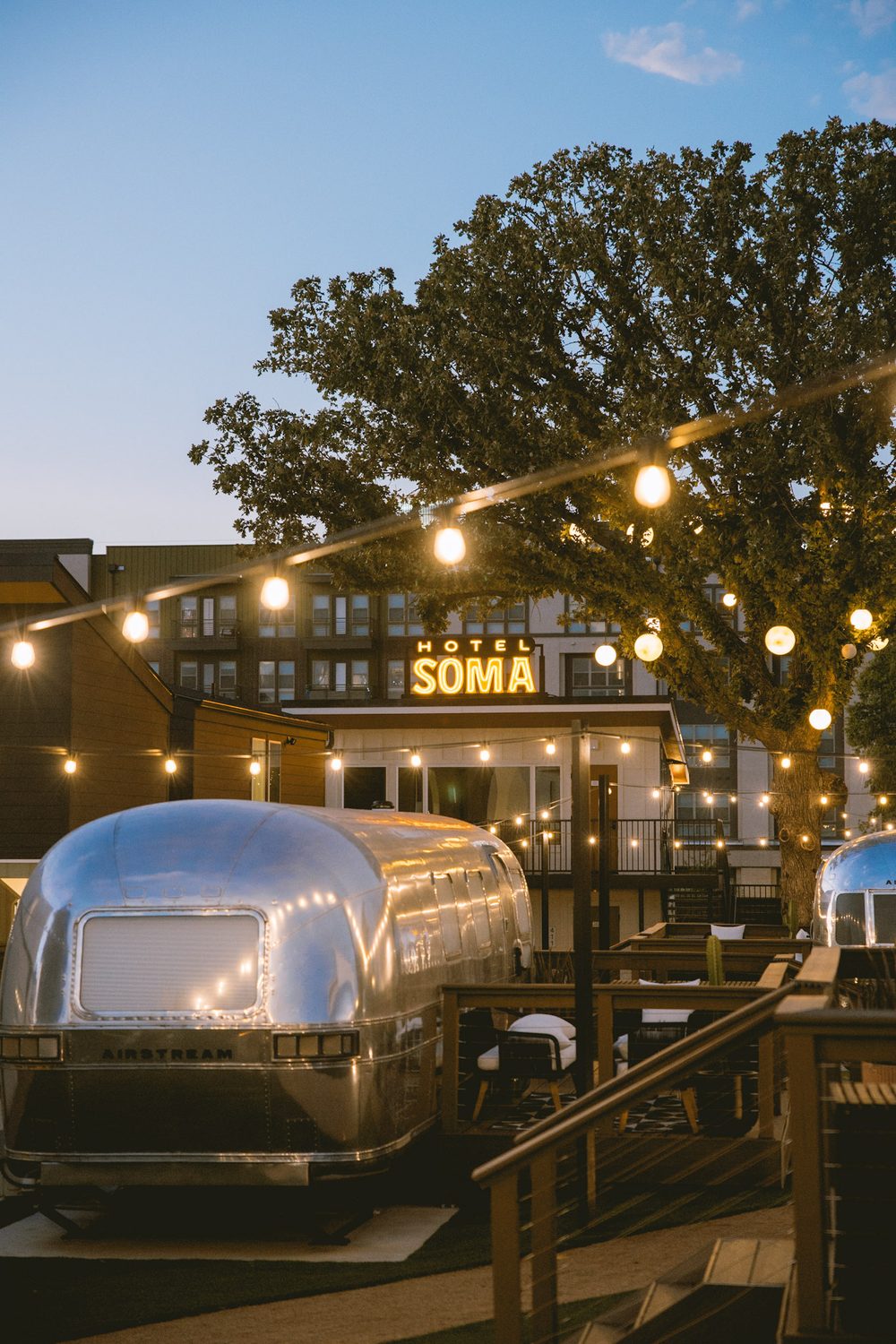Airstream suite at dusk under glowing string lights, with the lit Hotel SOMA sign in the background, Fort Worth
